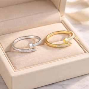 以下が含まれることがあります： ジュエリーボックスに入った2つの指輪。1つの指輪はシルバー色、もう1つは金色です。どちらの指輪も、小さな正方形の頭と、釘のシャフトに沿って小さな透明な石の列が付いた、曲がった釘のようにデザインされています。