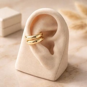 May include: Gold-coloured ear cuffs displayed on a beige, ear-shaped jewellery holder. The ear cuffs have a smooth, rounded design. A small, white jewellery box is visible in the background.
