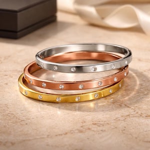 May include: Three metal bangles in silver, rose gold, and gold, each adorned with small, sparkling gemstones. The bangles are stacked, showcasing their different colors and textures. The jewelry is displayed on a light-colored surface.