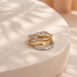 Puede incluir: Una pila de anillos en tonos dorados y plateados. Los anillos varían en diseño, algunos con un acabado liso y pulido y otros adornados con pequeños detalles brillantes. Los anillos están apilados sobre una superficie de tono neutro.