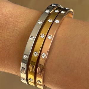 May include: Four metal bangles in silver, gold, and rose gold, each set with clear, round gemstones. The bracelets are stacked, displaying their different colours and designs. The bangles are on a wrist.