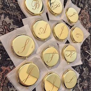 May include: A selection of gold-plated compact mirrors, each featuring a unique initial in a cursive script. The round mirrors are displayed on white tissue paper. The background is a patterned rug.
