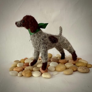 May include: A felted dog figurine in a standing pose, with a brown head and spots, gray body, and dark brown legs. It wears a green ribbon and collar, and stands on a bed of small, smooth stones. The dog's tail is upright.
