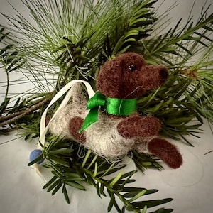May include: A brown and white felt dog ornament with a green ribbon around its neck. The ornament is hanging from a white string with a blue bead.