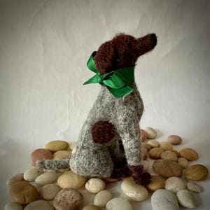 May include: A handcrafted felt dog figurine with a brown head and accents, a gray body, and a green ribbon. The dog is sitting on a bed of small, smooth stones. The figurine has a whimsical, artistic design.