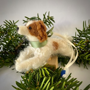 May include: A white and brown felt dog ornament with a green ribbon around its neck. The ornament is hanging from a white ribbon with a blue bead at the end.