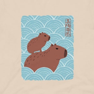 May include: A beige t-shirt with a graphic of two capybaras on a blue and white wave pattern. The Japanese text on the graphic reads "Capybara".