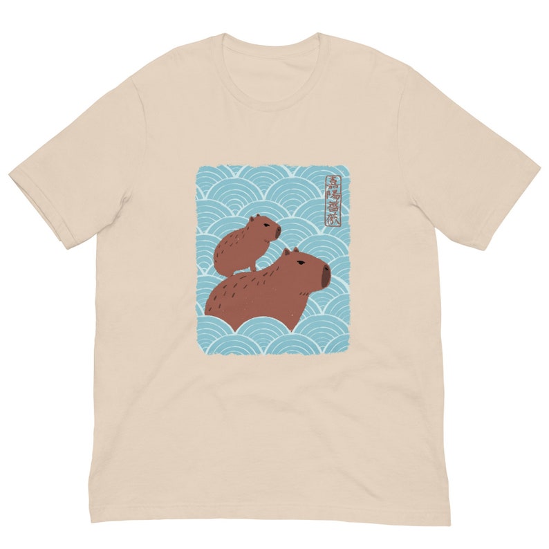 May include: A beige t-shirt with a graphic of two capybaras on a blue and white wave pattern. The Japanese text on the graphic reads "Capybara".