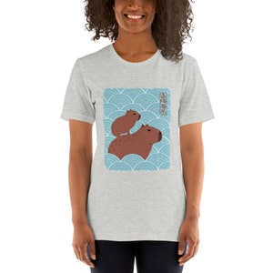 以下が含まれることがあります： 青と白の波模様に、2匹のカピバラが描かれたグレーのTシャツ。Tシャツに書かれた日本語は「カピバラ」です。