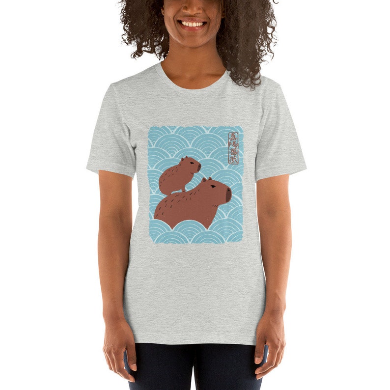 以下が含まれることがあります： 青と白の波模様に、2匹のカピバラが描かれたグレーのTシャツ。Tシャツに書かれた日本語は「カピバラ」です。