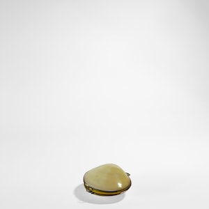 May include: A small, cream-coloured clam shell-shaped trinket box with a gold-coloured metal trim. The box is closed and sits on a white surface, casting a shadow. The shell has a smooth, glossy finish.