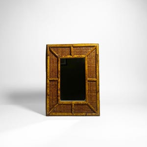 May include: Rectangular bamboo framed mirror. The frame has a woven texture and a warm, golden-brown color. The mirror's surface is dark, reflecting the surrounding environment. The design is simple and elegant, suitable for various decor styles.