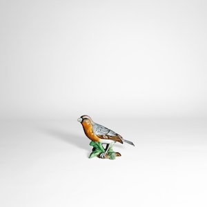 Könnte beinhalten: Eine Vogel-Figur aus Keramik mit detailliertem Design. Der Vogel hat einen grauen Kopf, eine orangefarbene Brust und grau-weiße Flügel. Er sitzt auf einem grünen Blattfuß. Die Figur ist ein dekorativer Artikel.