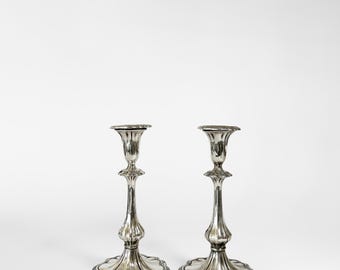 May include: Two silver candlesticks with ornate bases and slender stems. The candlesticks have a classic design with a scalloped edge on the base and a flared top to hold a candle. The silver finish gives them a vintage look.