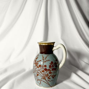 Könnte beinhalten: Eine blaue und braune Keramikkanne mit Blumenmuster. Die Kanne hat einen weißen Henkel und einen goldenen Rand.