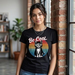 Peut inclure: Un t-shirt noir avec un motif central représentant un chat portant des lunettes de soleil à l'intérieur d'un cercle aux couleurs du coucher de soleil. Le texte "Be Cool" est imprimé en blanc au-dessus du chat. Col rond et manches courtes.