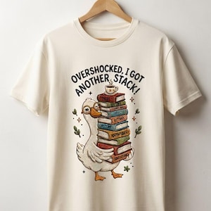 Peut inclure: Un t-shirt couleur crème avec un motif représentant un canard blanc portant des lunettes rondes et tenant une pile de livres colorés. Une petite tasse de café est posée sur les livres. Le texte indique, OVERSHOCKED, I GOT ANOTHER STACK!
