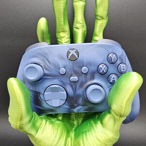 May include: A 3D-printed green hand holding a blue Xbox controller. The controller has the standard button layout with the Xbox logo. The hand is detailed with textured skin and is mounted on a circular base.