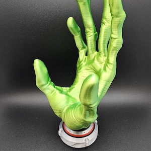 May include: A green, 3D-printed hand with long fingers, mounted on a circular base with a red accent. The hand is posed in a gesture, possibly a greeting or a display of support. The base is approximately 2 inches in diameter.