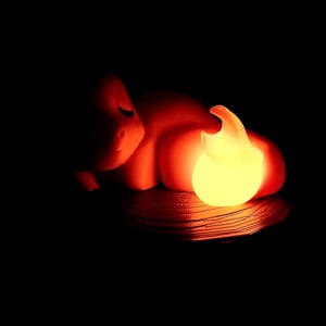May include: A glowing, flame-shaped night light in a dark setting. The light emits a warm, yellow glow, with orange and red hues. The base appears to be a woven material. The overall effect is a cozy, ambient light source.