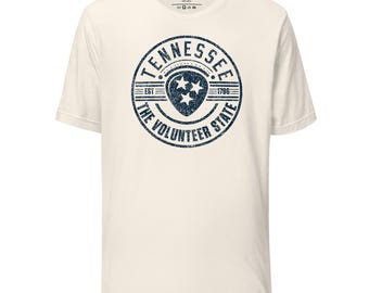 Könnte beinhalten: Ein weißes T-Shirt mit einer blauen, abgenutzten Grafik eines Kreises mit den Worten "Tennessee, The Volunteer State, Est 1796" und einem Gitarrenplektrum mit drei Sternen darin.