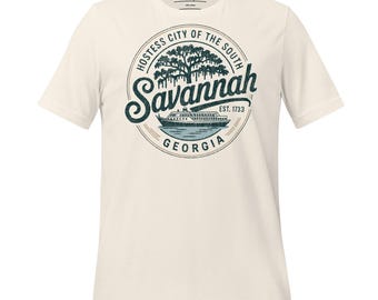 Puede incluir: Camiseta color crema con un diseño gráfico circular. El diseño incluye el texto "Savannah, Georgia", una ilustración de un árbol y un barco. También incluye el texto "Hostess City of the South" y "Est. 1733". La camiseta tiene mangas cortas y cuello redondo.