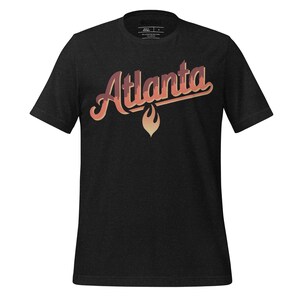 以下が含まれることがあります： グラデーションのオレンジと赤のスクリプトで「Atlanta」という文字が入った濃い灰色のTシャツ。テキストの下には様式化された炎のデザインがあります。シャツはクルーネックで半袖です。