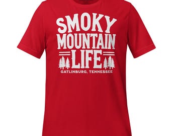以下が含まれることがあります： 白い文字で「Smoky Mountain Life」と書かれた赤いTシャツ。 「Life」の文字の上下に線があり、その上下に松の木が並んでいます。 木の下には「Gatlinburg, Tennessee」の文字があります。
