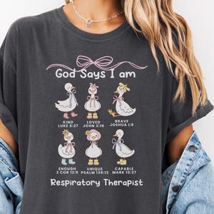May include: Dark gray t-shirt featuring a graphic of six cartoon ducks with the words "God Says I am" above them. Each duck has a different word associated with it, such as "Kind" and "Brave". The text "Respiratory Therapist" is printed at the bottom.