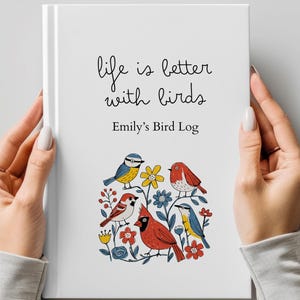 Può includere: Un libro con copertina rigida bianca intitolato Life is better with birds ed Emily's Bird Log, caratterizzato da un'illustrazione colorata di vari uccelli tra cui un cardinale rosso, una cinciarella e un passero circondati da fiori rossi, blu e gialli.