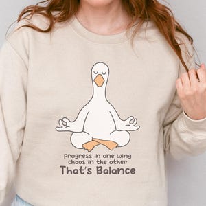 Op de afbeelding: Zandkleurige sweatshirt met een cartoon gans in een yogahouding. De gans is wit met oranje voeten en een gele snavel. De tekst "Progress in one wing chaos in the other, That's Balance" staat eronder.