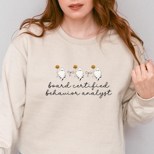 Op de afbeelding: Zandkleurige sweatshirt met de tekst "board certified behavior analyst" in zwart schrift. Het ontwerp bevat drie cartoongeesten met pompoenen. De sweatshirt heeft een neutrale kleur en een ronde hals.
