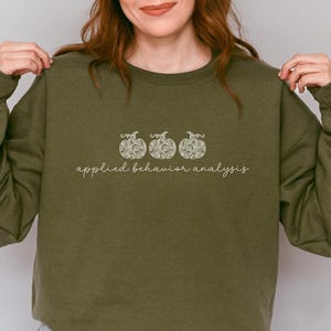 Può includere: Felpa verde oliva con la scritta "applied behavior analysis" e tre zucche decorative. La felpa è in tinta unita e ha lo scollo tondo. Il testo e le zucche sono di colore chiaro.