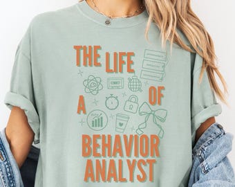 May include: A pale green t-shirt with the words "THE LIFE OF A BEHAVIOR ANALYST" in orange. The shirt has orange icons related to the profession. The model is wearing a light blue denim jacket.