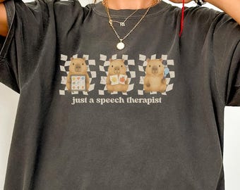 May include: Dark gray t-shirt with the text "Just a speech therapist." The shirt features a graphic of three cartoon animals on a checkered background. The animals are holding books and hearts.