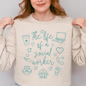 Puede incluir: Sudadera color arena con el texto "the life of a social worker" e iconos turquesas como una taza de café, un portátil y un teléfono. La sudadera es de cuello redondo.