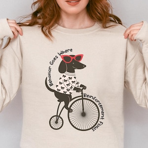Op de afbeelding: Zandkleurige sweatshirt met een teckel die een zonnebril en een trui draagt, op een fiets. De tekst "Behavior Goes Where Reinforcement Flows" staat rond de hond en de fiets gedrukt. De sweatshirt heeft lange mouwen.