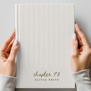 May include: A white hardback book with a subtle vertical striped pattern. The front cover features the text "chapter 70" in a gold cursive script font, followed by the name "OLIVIA SMITH" printed in a simple, dark, uppercase sans-serif font.
