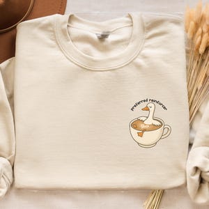 Op de afbeelding: Zandkleurige sweatshirt met een ronde hals. De sweatshirt heeft een afbeelding van een eend in een koffiekopje met de tekst "preferred reinforcer". Een bruine hoed en droogbloemen staan ook op de afbeelding.