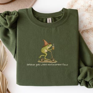 May include: Military green sweatshirt with a whimsical frog graphic. The frog, wearing a wizard hat, rides a scooter and holds a beverage. The text "behavior goes where reinforcement flows" is below the graphic. The sweatshirt is made of a soft material.