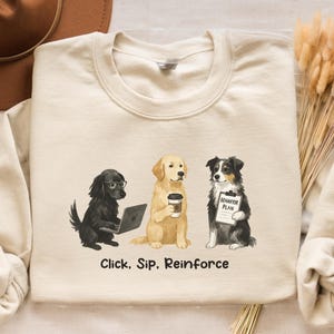 Op de afbeelding: Zandkleurige sweatshirt met een afbeelding van drie honden: een zwarte hond met een bril die een laptop gebruikt, een golden retriever met een koffiekopje en een zwart-witte hond met een "Behavior Plan". De tekst "Click, Sip, Reinforce" staat eronder.