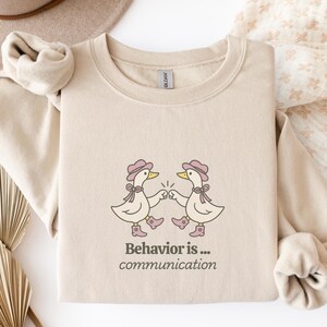 Op de afbeelding: Zandkleurige sweatshirt met een afbeelding van twee eenden met hoeden en laarzen, die hun vuisten tegen elkaar slaan. De tekst "Behavior is... communication" staat eronder. De sweatshirt heeft lange mouwen en een ronde hals.