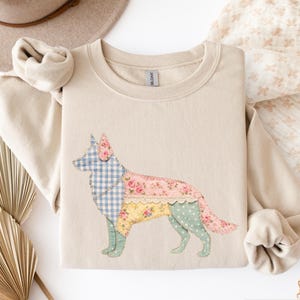 Puede incluir: Una sudadera de color arena con un diseño de perro de retazos. El perro presenta una cabeza de cuadros azules, un cuerpo floral y una parte inferior del cuerpo con estampado amarillo y verde. La sudadera tiene mangas largas y cuello redondo.