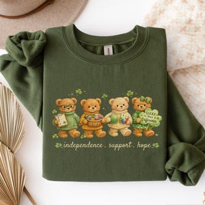 May include: Military green sweatshirt featuring a graphic of four teddy bears holding items and shamrocks. The text "independence. support. hope" is printed below the bears. The sweatshirt has a crew neck and long sleeves.