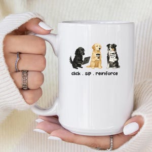 Könnte beinhalten: Weiße Keramiktasse mit einer Grafik von drei Hunden: einem schwarzen Hund mit einem Laptop, einem Golden Retriever mit Kaffee und einem Border Collie mit einem Schild. Der Text "click. sip. reinforce" steht darunter.