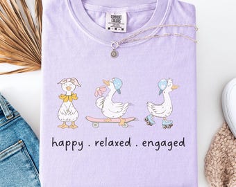 May include: A lilac Comfort Colors t-shirt featuring three cartoon ducks. One duck wears a bow, another rides a skateboard, and the third wears roller skates. The text "happy . relaxed . engaged" is below the ducks.