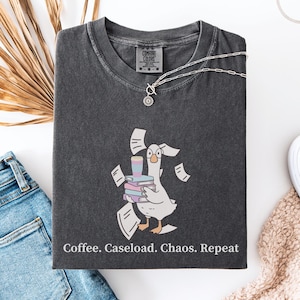 Peut inclure: T-shirt gris foncé avec un dessin d'oie portant des livres, une tasse de café et des papiers. Le texte "Coffee. Caseload. Chaos. Repeat" est imprimé en bas. Le t-shirt est associé à un jean bleu et des baskets blanches.