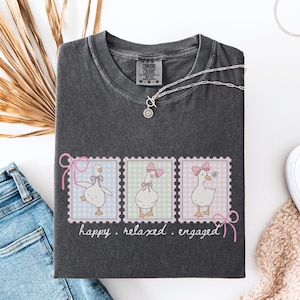 Peut inclure: T-shirt Comfort Colors gris foncé avec trois oies de dessin animé dans des motifs de timbres-poste. Chaque oie porte un nœud rose. Les mots "happy. relaxed. engaged." sont écrits sous les timbres. Un collier argenté et des baskets blanches sont également visibles.