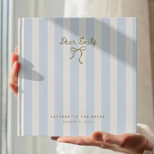 Può includere: Un libro quadrato con una copertina a righe bianche e azzurre. Il testo "Dear Emily" e un fiocco dorato. Sotto, il testo recita "Letters to the Bride August 9, 2026". Il libro è tenuto davanti a una finestra.