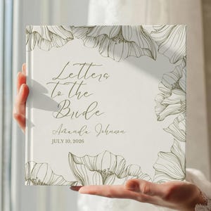 May include: A square, off-white book titled "Letters to the Bride" with floral illustrations. The cover includes the name "Amanda Johnson" and the date "July 10, 2026" in elegant script. The book is held in front of a soft, blurred background.